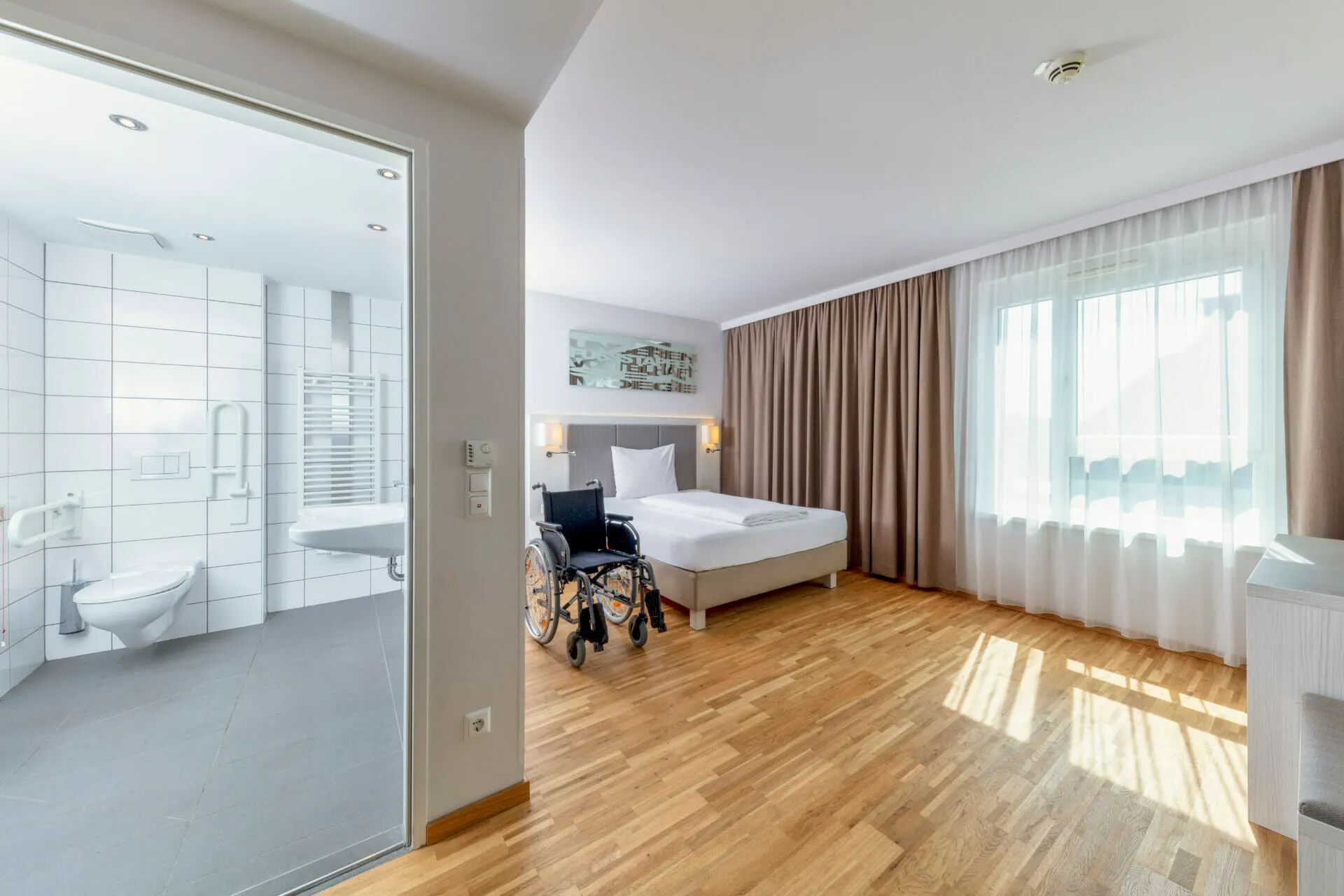 Barrierefreies Zimmer mit Rollstuhl und Blick durch die Glaswand ins Bad
