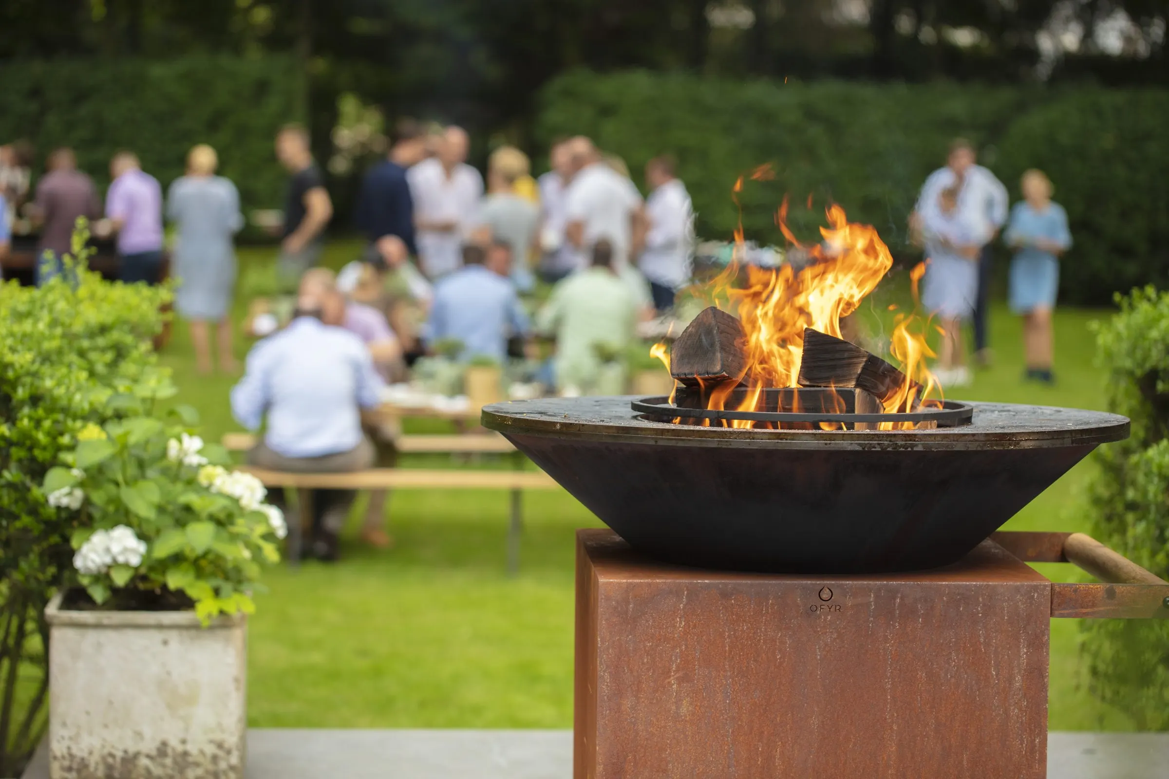 Gäste stehen und sitzen im Garten, davor ein OFYR mit lodernden Holzscheiten