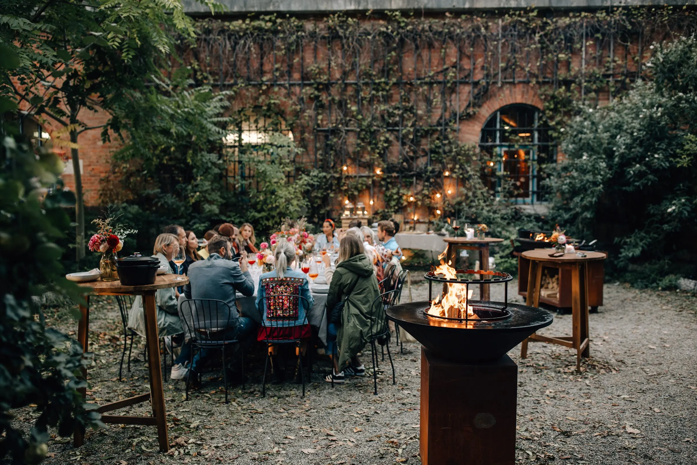 Gedeckte Tafel im Innenhof vor einer Ziegelmauer, daneben ein OFYR mit Feuer in der Dämmerung
