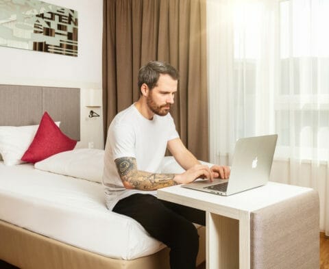 Gast sitzt am Bett und arbeitet am Laptop. Der umgestellte Kofferbock dient als Schreibtisch.